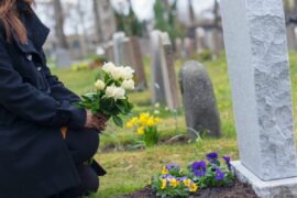 An adult in a black coat holds flowers in front of a newly dug grave in a cemetery