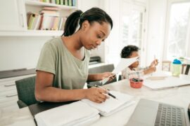 An adult looks at stack of papers in a folder in front of a laptop computer, a young child drawing next to her