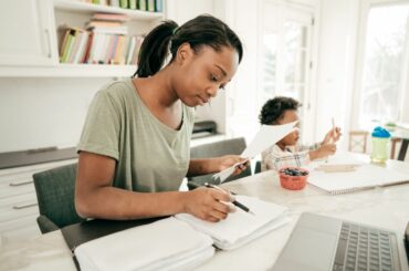 An adult looks at stack of papers in a folder in front of a laptop computer, a young child drawing next to her