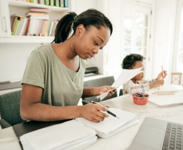 An adult looks at stack of papers in a folder in front of a laptop computer, a young child drawing next to her