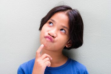 An Asian child looks up with a confused expression and a finger on their chin