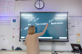 A teacher writes on a digital smart board at the front of a classroom