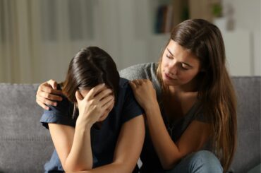 Teenager with long hair puts her arm around a friend who is covering her face in distress