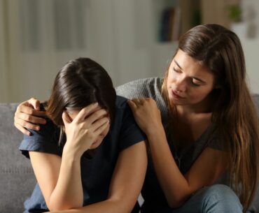 Teenager with long hair puts her arm around a friend who is covering her face in distress