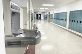 A water drinking fountain in a hallway lined with lockers and fluorescent lighting