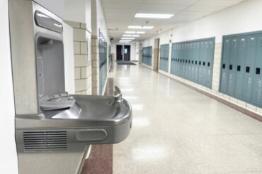A water drinking fountain in a hallway lined with lockers and fluorescent lighting