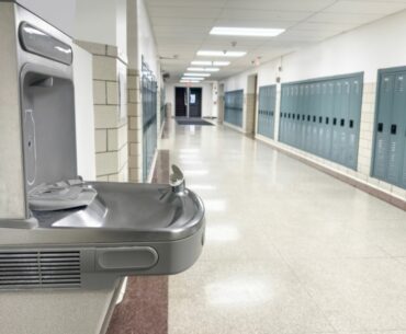 A water drinking fountain in a hallway lined with lockers and fluorescent lighting