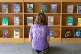 An adolescent in a purple t-shirt kneels on the carpet in front of a library shelf of books