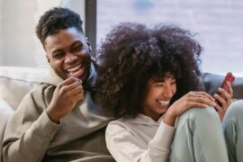 A young Black couple in sweats, one of them holding a phone, laughs together on the couch