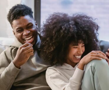 A young Black couple in sweats, one of them holding a phone, laughs together on the couch