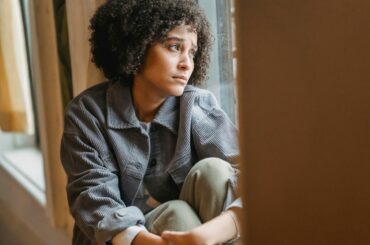 Woman with curly black hair, brown skin and a sad facial expression looking out the window