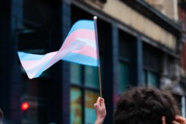 A hand holds a trans pride flag outside of a building