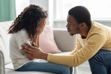 A child covers her face on the couch, while a dad comforts her