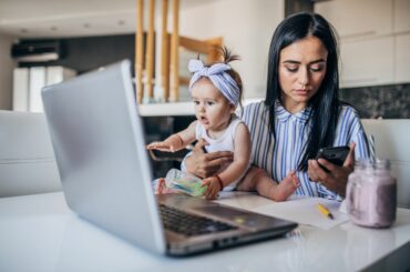 A stressed mom with baby looking at phone in front of a laptop computer