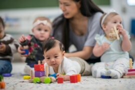 Four babies explore toys on the rug at a day care center, while a teacher helps one of the babies
