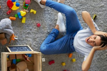 A stressed parent yells, head in hands, while sitting on a rug surrounded by messy toys