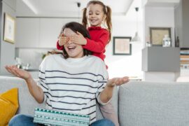 A child stands behind mother, covering her eyes to surprise her with a gift