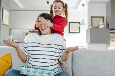 A child stands behind mother, covering her eyes to surprise her with a gift