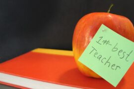 An apple sits on top of a book, and a green post-it note on the apple reads "#1 best teacher" in a child's handwriting