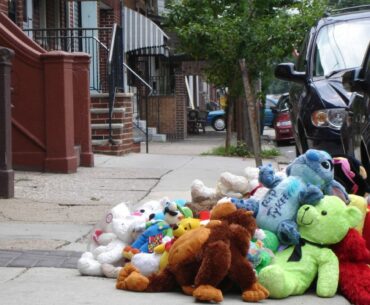In a large pile of stuffed animals on a city sidewalk, a blue Stitch character on top reads in marker, "R.I.P. Tykeem" for a victim of gun violence.