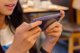 A teen or tween with long dark brown hair holds a smartphone horizontally in a purple case