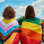 two people with backs turned with Pride flags draped over their shoulders