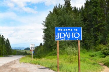 A blue sign reads "Welcome to Idaho" next to a hilly road, evergreen trees, and a view of mountains in the background