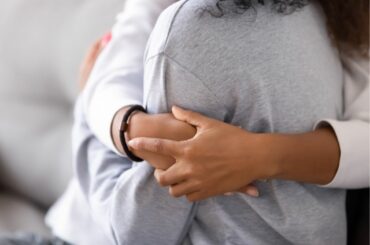A closeup of a teenager and an adult hugging. Both have brown skin and light gray shirts.
