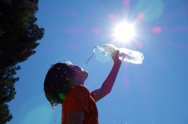 A child leans head back and pours water into mouth, under a hot sun with no clouds