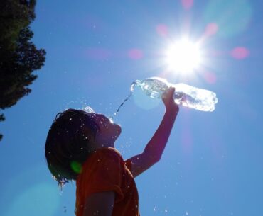 A child leans head back and pours water into mouth, under a hot sun with no clouds