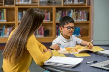 Young brown-haired child with glasses gets help from a tutor at a table in the library