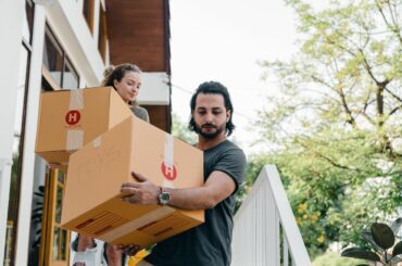 Two people carry cardboard boxes down the steps outside of a home