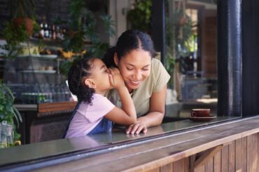 A young child cups hand to her parent's ear to whisper a secret in the window of a coffee shop