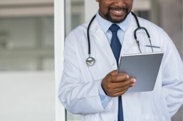 A doctor with a beard and brown skin wears a white coat with a stethoscope draped over shoulders and smiles as he looks down at a tablet