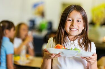Child smiles in school cafeteria, holding up a white tray with food that appears to include fruits and vegetables