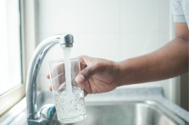 An adult hand with brown skin holds a drinking glass under a kitchen water faucet
