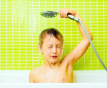 A light-skinned boy holds a shower head over his head, eyes closed, in front of a bright green tile wall
