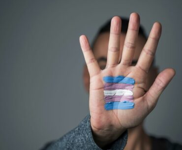 A person's hand up in front of their face, with a blue, pink, and white trans flag painted on the palm of their hand