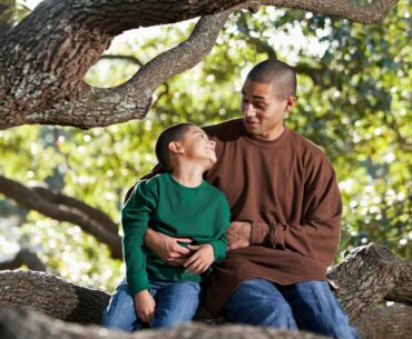 A dad and son with brown skin and short black hair talk and smile at each other while sitting on a large tree branch