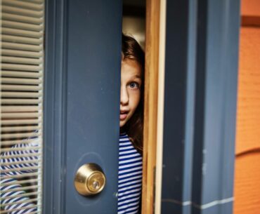 A white child with long hair and striped shirt opens the door of a house and peeks outside, a wide-eyed expression on her face