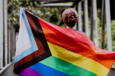 A view of the back of a person with short curly hair holding a Pride Progress flag stretched over their back and arms, as they walk over a pedestrian bridge
