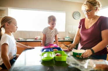 A mom packs three lunch boxes while two kids look on in the kitchen