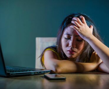 A young woman or teen girl clutches her head, looking scared or distressed next to a phone and laptop