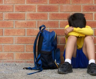 A dark-haired child sits against a brick wall with their head hidden in their arms. Muslim and Jewish children have been experiencing bullying.