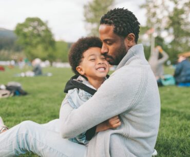 A young kid and dad share a warm embrace while sitting on grass in a park
