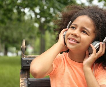 Happy young kid with afro hairstyle listens to music on headphones while sitting on a park bench