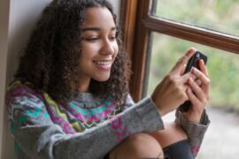 A smiling teen looks at the phone in her hands while sitting against a wall.