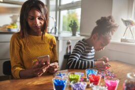 A parent using AI productivity tools on her phone while a child does colorful bead crafts beside her