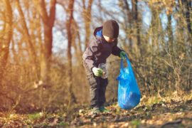 A child wearing a winter hat and work gloves picks up garbage in a wooded area