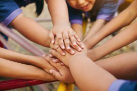 A group of kids with different shades of brown skin stack their hands together on a playground